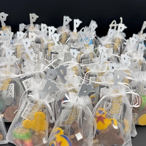 Puede incluir: Una colección de pequeñas bolsas de organza transparentes llenas de caramelos de colores y con formas. Cada bolsa está atada con una cinta blanca y rematada con una decoración plateada en forma de bandera. Algunas bolsas tienen etiquetas con códigos QR.