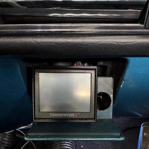Puede incluir: Una pantalla digital rectangular negra con el texto TERMINATOR X impreso en blanco en el marco inferior. El dispositivo está montado dentro de un panel de salpicadero de coche color verde azulado, con un recorte circular en el lado derecho.