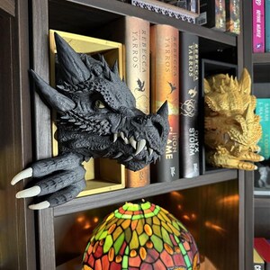 May include: A dark gray dragon head bookshelf decor with sharp teeth and claws, emerging from a wooden shelf. A stained glass lamp with dragonfly design sits below. Books with titles like "Rebecca Yarros" and "Iron Flame" are visible.