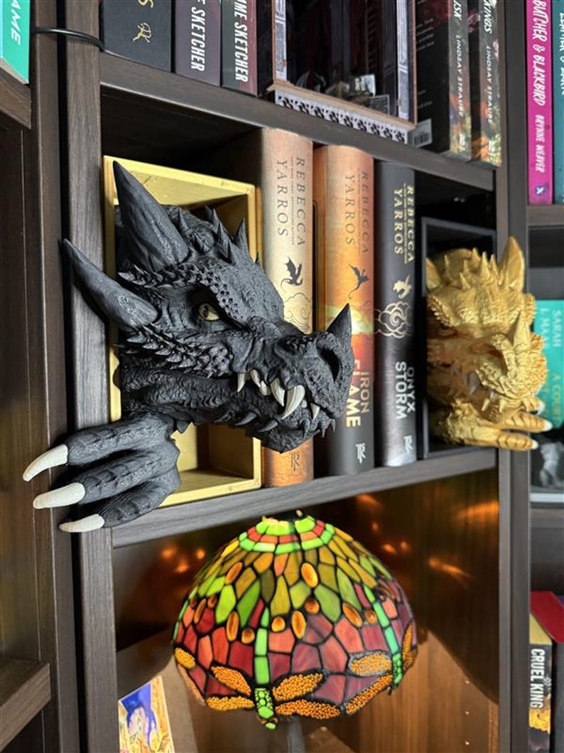 May include: A dark gray dragon head bookshelf decor with sharp teeth and claws, emerging from a wooden shelf. A stained glass lamp with dragonfly design sits below. Books with titles like "Rebecca Yarros" and "Iron Flame" are visible.