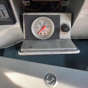 May include: An Auto Meter Pro-Comp Ultra-Lite fuel pressure gauge with a white face and red needle, mounted in a rectangular housing with a black textured panel next to a cigarette lighter, installed within a silver automotive dashboard console.