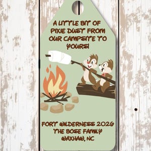 May include: A light green rectangular tag with a hole at the top, featuring two cartoon chipmunks roasting a marshmallow over a campfire. Text reads: A LITTLE BIT OF PIXIE DUST FROM OUR CAMPSITE TO YOURS! FORT WILDERNESS 2026 THE BOSE FAMILY WAXHAW, NC.