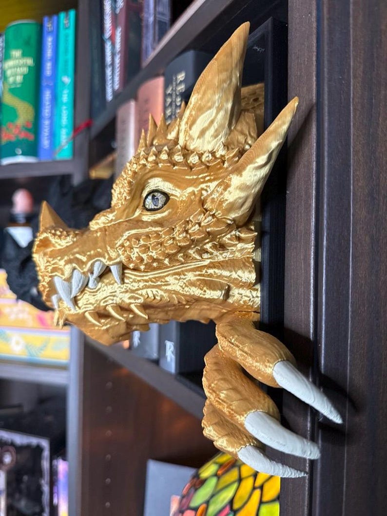 May include: A gold-colored dragon head and claw bookshelf decor. The dragon head has detailed scales, horns, and sharp teeth. The claw extends from the side, adding a unique touch to any bookshelf. The dragon's eye is a light blue color.
