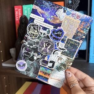 May include: A rectangular holographic sticker sheet featuring various fantasy-themed illustrations, including hands reaching for stars, a Velaris postage stamp, and text reading "to the stars who listen" and "High Lady of the Smut Court" in dark tones.