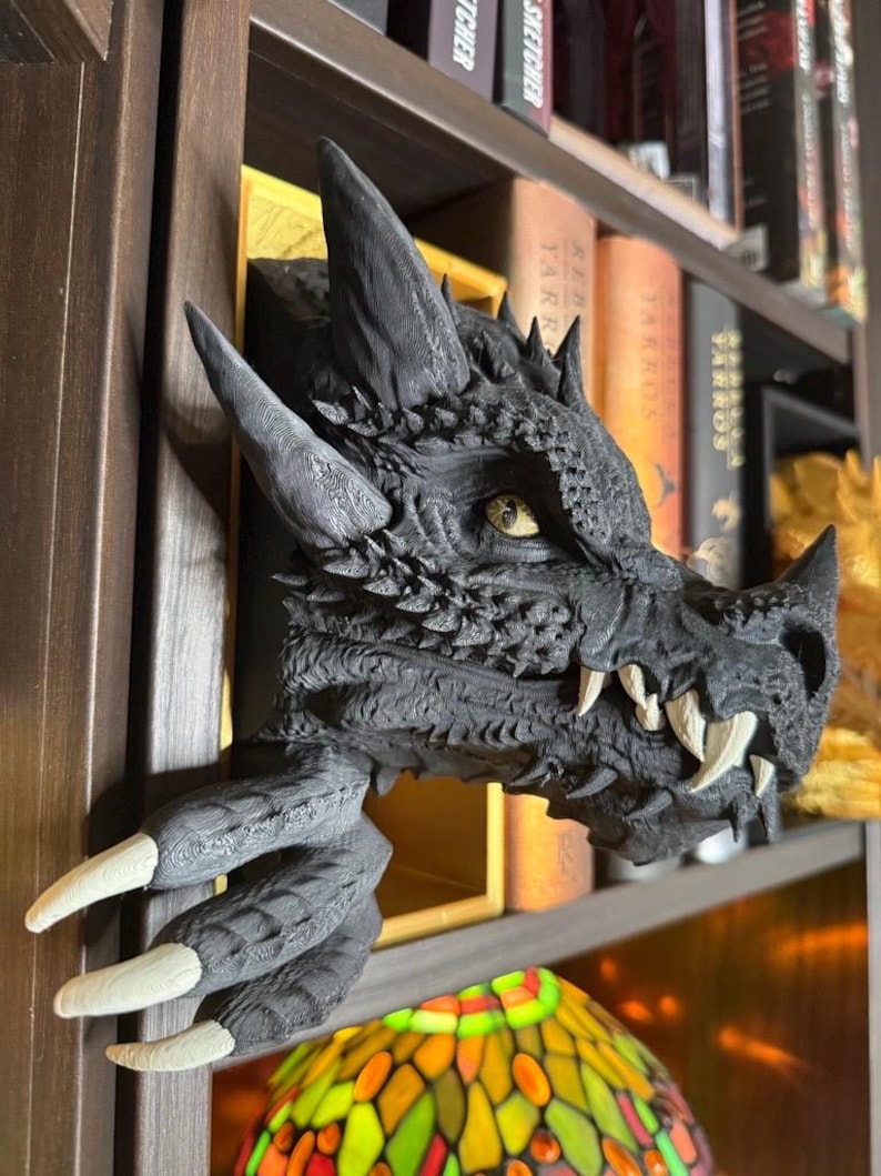 May include: A detailed, black dragon head sculpture with sharp horns, textured scales, and prominent white fangs. The dragon's claw extends from the bookshelf, with long, white claws. The sculpture is a decorative item for fantasy enthusiasts.