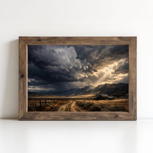 May include: A framed landscape print featuring a dramatic sky with dark storm clouds over a rural scene. A dirt road leads to a small building near the mountains. The frame is made of rustic wood.