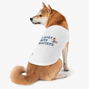 Pode incluir: Um cão Shiba Inu vestindo uma camiseta branca com o texto "I ONLY BITE RACISTS" em azul. A camiseta tem um pequeno gráfico de uma pessoa fugindo de um cachorro. O cão é marrom e branco.