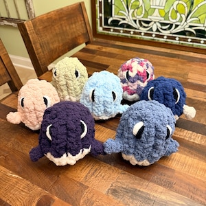 May include: A collection of handmade crochet octopus toys in various colors, including pink, light blue, and purple. Each octopus has white and black eyes and small tentacles. The toys are arranged on a wooden table.