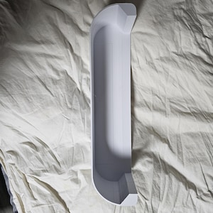 May include: A white, elongated, curved object with a rectangular channel. The object has a geometric design at each end. The item is resting on a wrinkled, light-colored fabric.