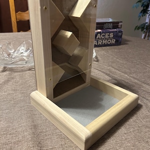 May include: A wooden dice tower with a clear acrylic front panel, featuring geometric shapes inside. The tower has a tray at the base lined with gray felt. The construction is light-colored wood, with brass-colored screws.
