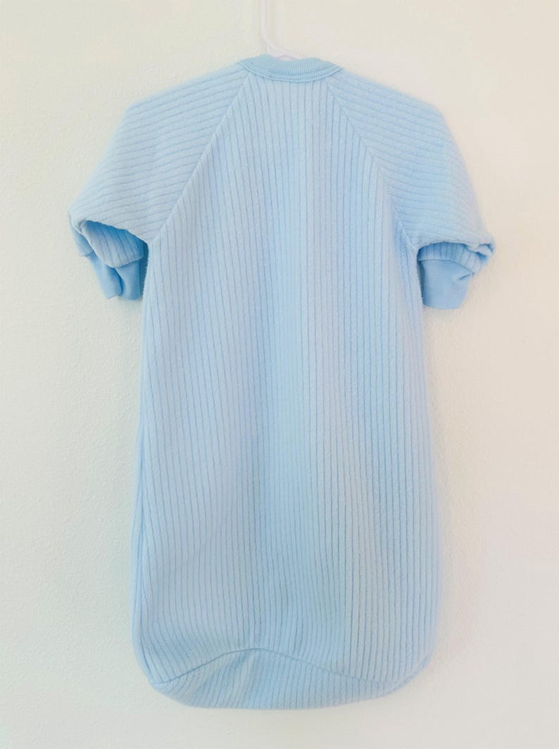 Puede incluir: Saco de dormir para beb&eacute; azul claro con mangas cortas, colgado de una percha blanca. El saco de dormir tiene una textura acanalada y una parte inferior redondeada. El saco de dormir est&aacute; hecho de un material suave.