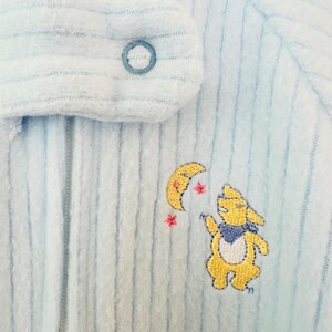 Puede incluir: Pijama de beb&eacute; azul claro con textura acanalada. El pijama tiene un cierre de bot&oacute;n y un dise&ntilde;o bordado de un conejito amarillo, una luna creciente y estrellas. La palabra "class" es visible en el cuello.