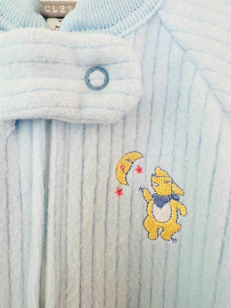 Puede incluir: Pijama de beb&eacute; azul claro con textura acanalada. El pijama tiene un cierre de bot&oacute;n y un dise&ntilde;o bordado de un conejito amarillo, una luna creciente y estrellas. La palabra "class" es visible en el cuello.