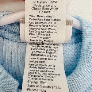 Puede incluir: Primer plano de una etiqueta de ropa con instrucciones de cuidado. La etiqueta es blanca con texto negro y est&aacute; adherida a una prenda azul claro. La etiqueta incluye instrucciones de lavado, como lavar a m&aacute;quina con agua tibia y secar en secadora a temperatura media.