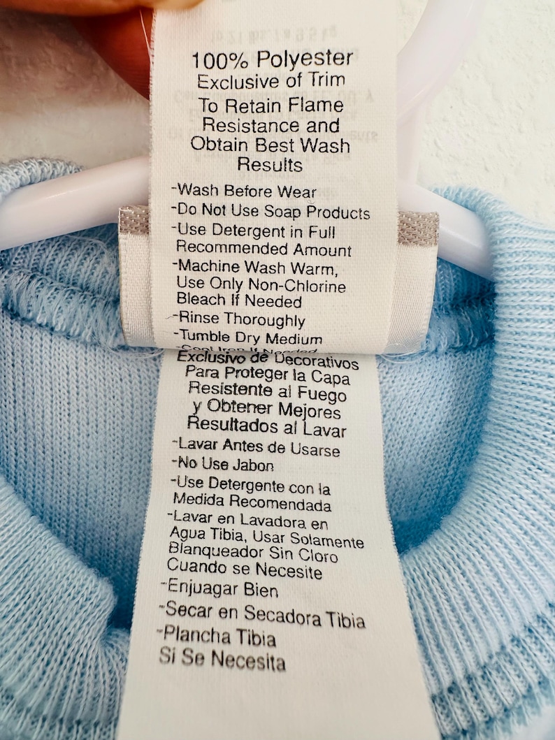 Puede incluir: Primer plano de una etiqueta de ropa con instrucciones de cuidado. La etiqueta es blanca con texto negro y est&aacute; adherida a una prenda azul claro. La etiqueta incluye instrucciones de lavado, como lavar a m&aacute;quina con agua tibia y secar en secadora a temperatura media.