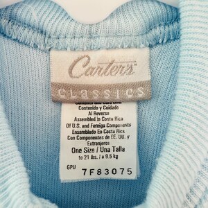 Puede incluir: Un su&eacute;ter de beb&eacute; azul claro con cremallera y una etiqueta que dice "Carters Classics". La etiqueta indica que es talla &uacute;nica, hasta 9,5 kg. El su&eacute;ter est&aacute; colgado en una percha blanca.