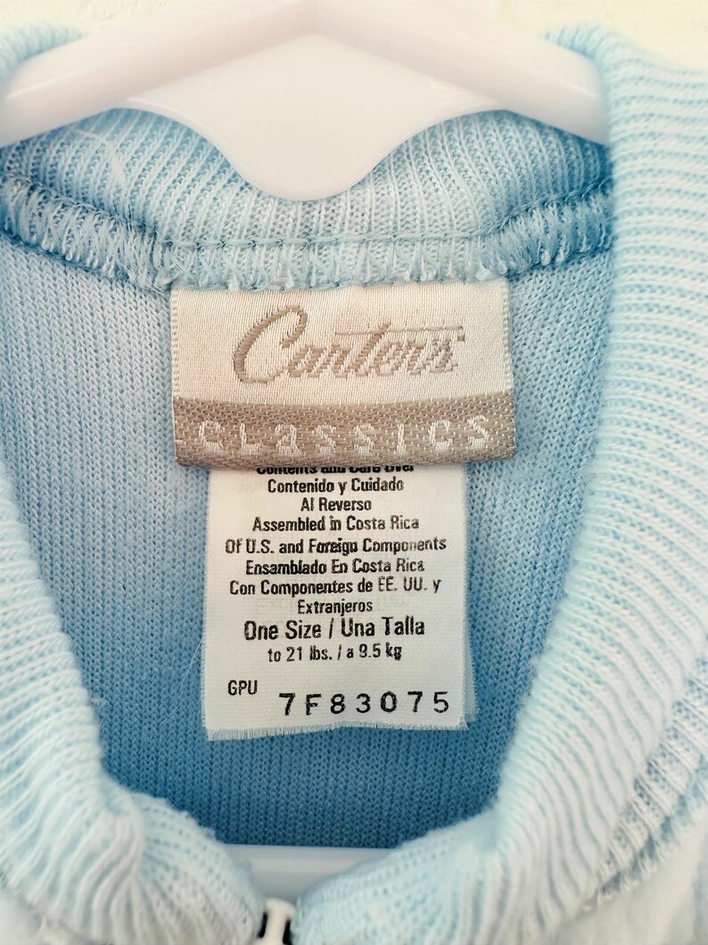 Puede incluir: Un su&eacute;ter de beb&eacute; azul claro con cremallera y una etiqueta que dice "Carters Classics". La etiqueta indica que es talla &uacute;nica, hasta 9,5 kg. El su&eacute;ter est&aacute; colgado en una percha blanca.