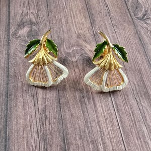 May include: A pair of gold-tone metal earrings shaped like bell-shaped flowers. Each earring features a textured gold stem, a bright green enamelled leaf, and white enamelled petals with thin gold-tone wire detailing radiating from the centre.