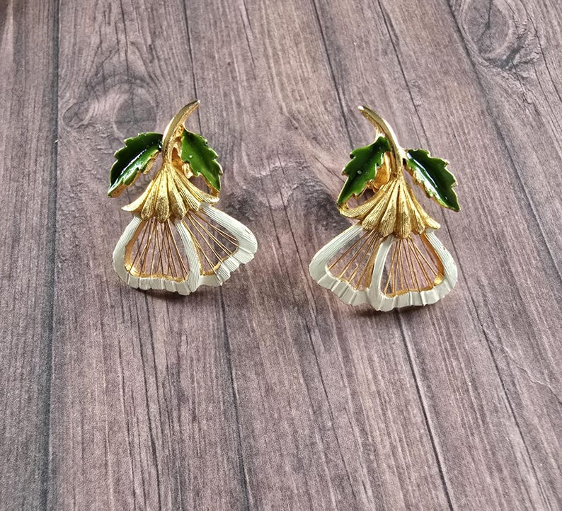 May include: A pair of gold-tone metal earrings shaped like bell-shaped flowers. Each earring features a textured gold stem, a bright green enamelled leaf, and white enamelled petals with thin gold-tone wire detailing radiating from the centre.