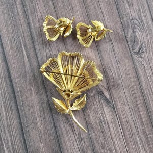 May include: A gold-tone metal jewellery set consisting of a large flower brooch and two matching clip-on earrings. The pieces feature a textured, wire-like petal design with a stem and leaf detail on the brooch, showing the back pin mechanism.