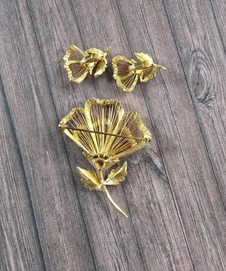May include: A gold-tone metal jewellery set consisting of a large flower brooch and two matching clip-on earrings. The pieces feature a textured, wire-like petal design with a stem and leaf detail on the brooch, showing the back pin mechanism.