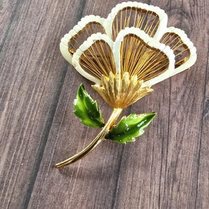 May include: A gold-tone metal flower brooch featuring white enamel-coated petals with thin gold wire detailing inside. The stem and two leaves are finished with a glossy green enamel coating, creating a vibrant contrast against the metallic gold base.