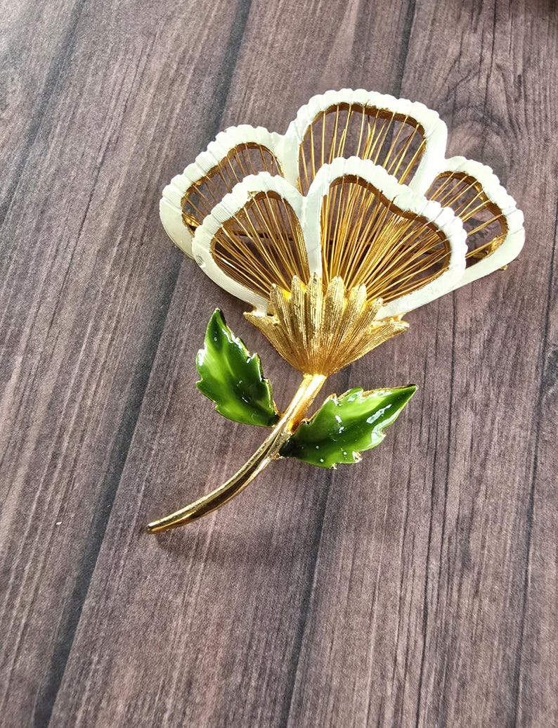 May include: A gold-tone metal flower brooch featuring white enamel-coated petals with thin gold wire detailing inside. The stem and two leaves are finished with a glossy green enamel coating, creating a vibrant contrast against the metallic gold base.