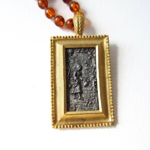 Puede incluir: Un collar con colgante rectangular dorado con una escena en relieve detallada. El colgante está enmarcado con un borde de cuentas y cuelga de un collar de cuentas de color ámbar. El relieve representa una figura en un jardín.