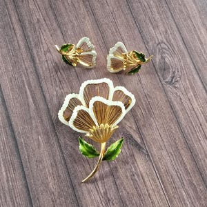 May include: A gold-tone metal jewellery set consisting of a large flower brooch and a pair of matching flower earrings. Each piece features white enamel-edged petals with fine gold wire detailing and vibrant green enamel leaves on the stems.