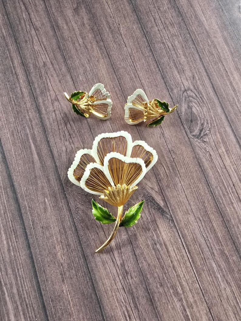 May include: A gold-tone metal jewellery set consisting of a large flower brooch and a pair of matching flower earrings. Each piece features white enamel-edged petals with fine gold wire detailing and vibrant green enamel leaves on the stems.