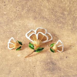 May include: A gold-tone metal jewellery set consisting of a large flower brooch and two matching smaller flower earrings. Each piece features white enamel petal edges, textured gold-tone centres, and vibrant green enamel leaves on the stems.