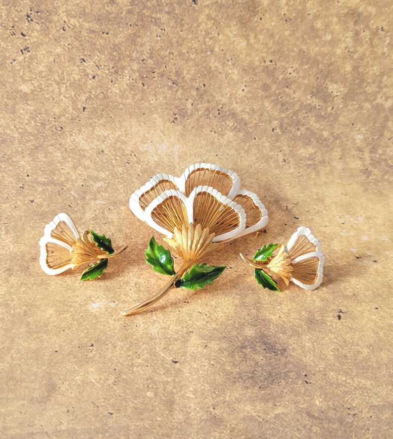 May include: A gold-tone metal jewellery set consisting of a large flower brooch and two matching smaller flower earrings. Each piece features white enamel petal edges, textured gold-tone centres, and vibrant green enamel leaves on the stems.