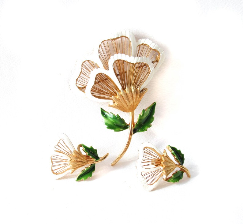 May include: A gold-tone metal jewellery set consisting of a large flower brooch and two matching smaller flower earrings. Each piece features white enamel petal tips, gold wire-work detailing, and vibrant green enameled leaves on the stems.