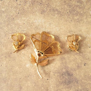 May include: A gold-tone metal jewellery set consisting of one large flower brooch and two matching flower earrings. The pieces feature textured, wire-like petal details and stems. The brooch includes a horizontal pin mechanism on the back.