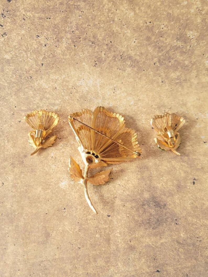 May include: A gold-tone metal jewellery set consisting of one large flower brooch and two matching flower earrings. The pieces feature textured, wire-like petal details and stems. The brooch includes a horizontal pin mechanism on the back.