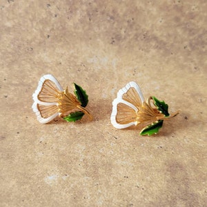 May include: A pair of gold-tone metal earrings shaped like flowers. Each earring features white enamel petals with fine gold-tone wire detailing, a textured gold-tone centre, and a small green enamel leaf attached to the stem of the flower.