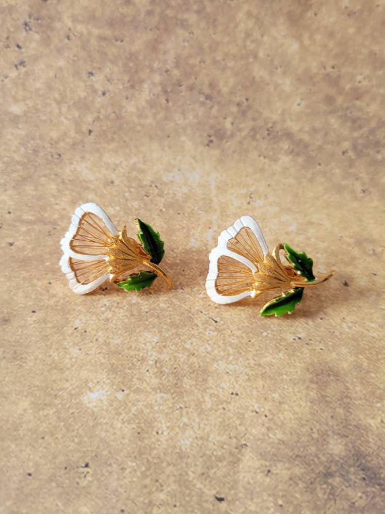 May include: A pair of gold-tone metal earrings shaped like flowers. Each earring features white enamel petals with fine gold-tone wire detailing, a textured gold-tone centre, and a small green enamel leaf attached to the stem of the flower.