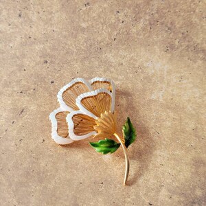 May include: A gold-tone metal flower brooch featuring three layered petals with white enamel edges and fine gold wire detailing. The flower has a textured gold centre and a smooth gold stem adorned with two vibrant green enamel leaves.