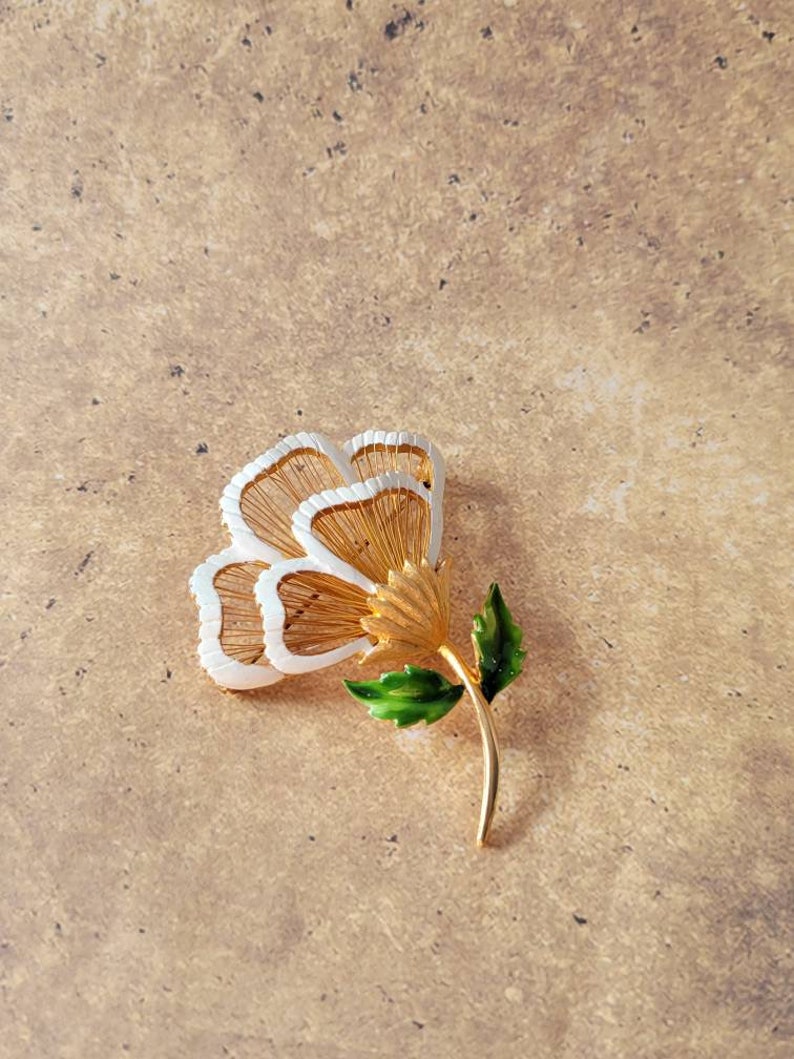 May include: A gold-tone metal flower brooch featuring three layered petals with white enamel edges and fine gold wire detailing. The flower has a textured gold centre and a smooth gold stem adorned with two vibrant green enamel leaves.