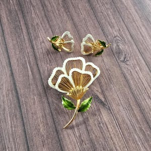 May include: A gold-tone jewellery set consisting of a large flower brooch and a pair of matching flower earrings. Each piece features white enamel petals with gold-tone wire detailing and vibrant green enamel leaves on the stems and bases.