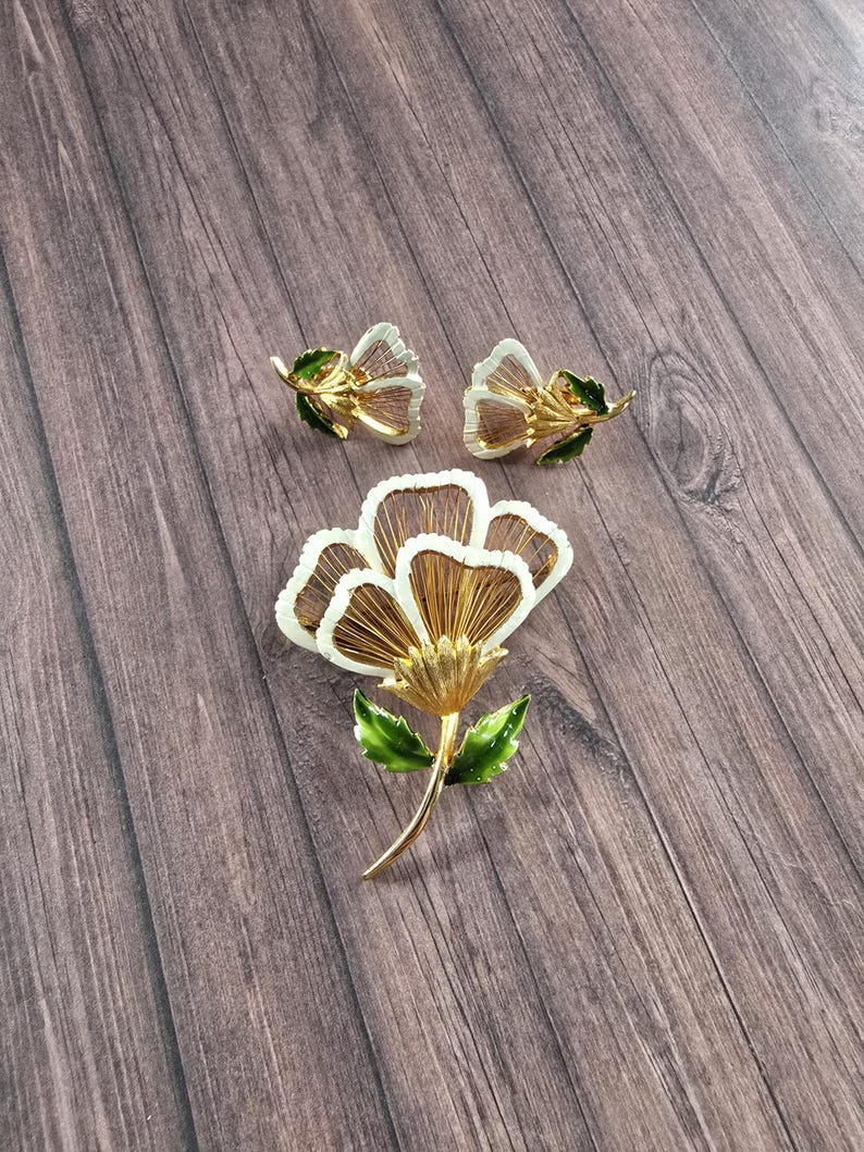 May include: A gold-tone jewellery set consisting of a large flower brooch and a pair of matching flower earrings. Each piece features white enamel petals with gold-tone wire detailing and vibrant green enamel leaves on the stems and bases.