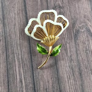 May include: A gold-tone metal brooch shaped like a flower with three large petals featuring white enamel edges and thin gold wire filaments inside. The flower has a textured gold centre and a stem with two glossy green enamel leaves attached.