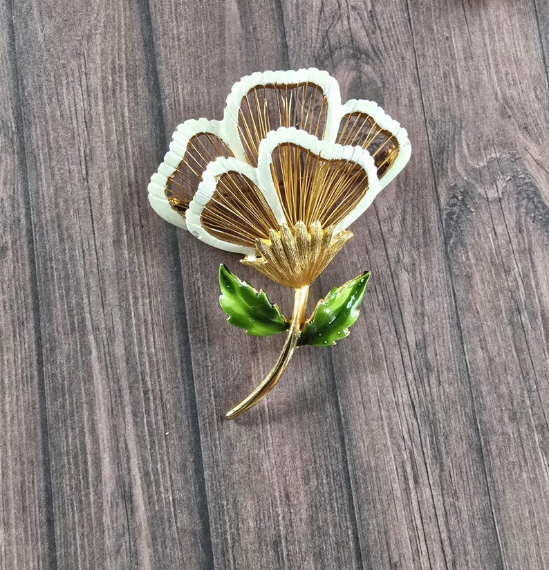 May include: A gold-tone metal brooch shaped like a flower with three large petals featuring white enamel edges and thin gold wire filaments inside. The flower has a textured gold centre and a stem with two glossy green enamel leaves attached.