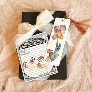 May include: A black book tied with a cream ribbon, alongside a watercolour bookmark depicting a purple iris and a card with the text "Archive Impressions". Copper-coloured paper clips are scattered around the card.