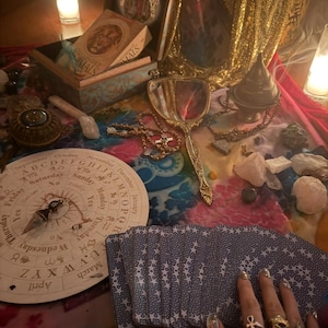 Puede incluir: Una colección de objetos místicos sobre una superficie colorida. Incluye una baraja de cartas con estrellas blancas, una tabla de péndulo, un espejo de mano, una caja con un libro y un rosario. La escena está iluminada por la luz de las velas, creando un ambiente cálido.