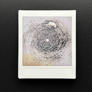 May include: A square Polaroid photograph featuring an abstract design. The image showcases a central white dot surrounded by a swirling pattern of black and gray marks, resembling a vortex or spiral. The photograph is set against a black background.