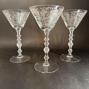 May include: Three clear, stemmed cocktail glasses with etched floral designs. The glasses have a conical bowl, a decorative stem with a beaded section, and a round base. The glasses are arranged on a dark surface.