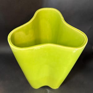 May include: A bright lime green ceramic vase with a unique, wavy top edge and a flared, angular shape. The vase has a glossy finish and a modern, minimalist design. The vase is suitable for displaying flowers or as a decorative accent.