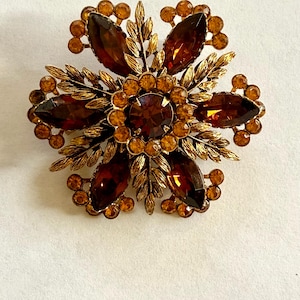 May include: A vintage brooch in a floral design. The brooch features large, faceted, amber-colored stones set in a gold-toned metal. The design includes leaf and berry accents, creating a dimensional and elegant accessory.