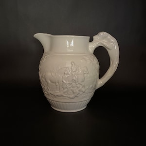 May include: White ceramic pitcher with a detailed relief design. The pitcher features a spout, a handle with a sculpted animal head, and a decorative band around the base. The relief depicts figures and animals. The background is black.